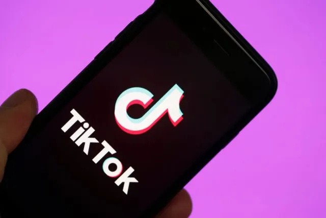 picture of tik tok app on phone held in a hand with a pink background